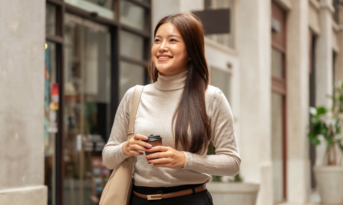 Smiling woman in a cream jumper walking down a city street holding a coffee