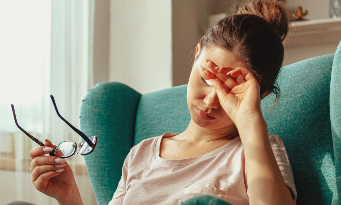 Woman in an armchair rubbing her eye with one hand and holding her glasses in the other